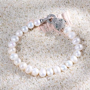Può includere: Un bracciale di perle con chiusura argentata a forma di testa di pantera. Il bracciale è composto da numerose perle bianche di forma ovale. La chiusura a testa di pantera è decorata con piccole pietre trasparenti e dettagli verdi.