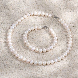 Puede incluir: Un collar de perlas blancas y un juego de pulsera a juego. Las joyas se exhiben sobre una superficie arenosa, con el collar formando una forma de espiral. Las perlas son redondas y tienen un brillo lustroso, y los cierres son plateados.