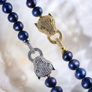 Può includere: Due collane con perle blu scuro. Una collana ha una chiusura a testa di pantera argentata con occhi verdi, l'altra una chiusura a testa di pantera dorata con occhi verdi. Le collane sono su una superficie bianca.