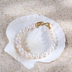 Puede incluir: Una pulsera de perlas con un cierre dorado descansa dentro de una concha blanca sobre una superficie arenosa. La pulsera está compuesta por numerosas perlas blancas redondas. El cierre dorado tiene un diseño delicado y curvado. La escena está iluminada con luz natural suave.