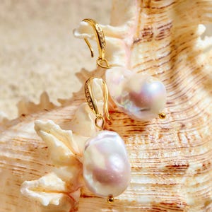 Puede incluir: Pendientes dorados con perlas grandes de forma irregular. Los pendientes tienen un diseño de gancho con pequeños adornos brillantes. Las perlas muestran un brillo suave e iridiscente, con toques de rosa y crema. Se encuentran sobre una concha beige texturizada.