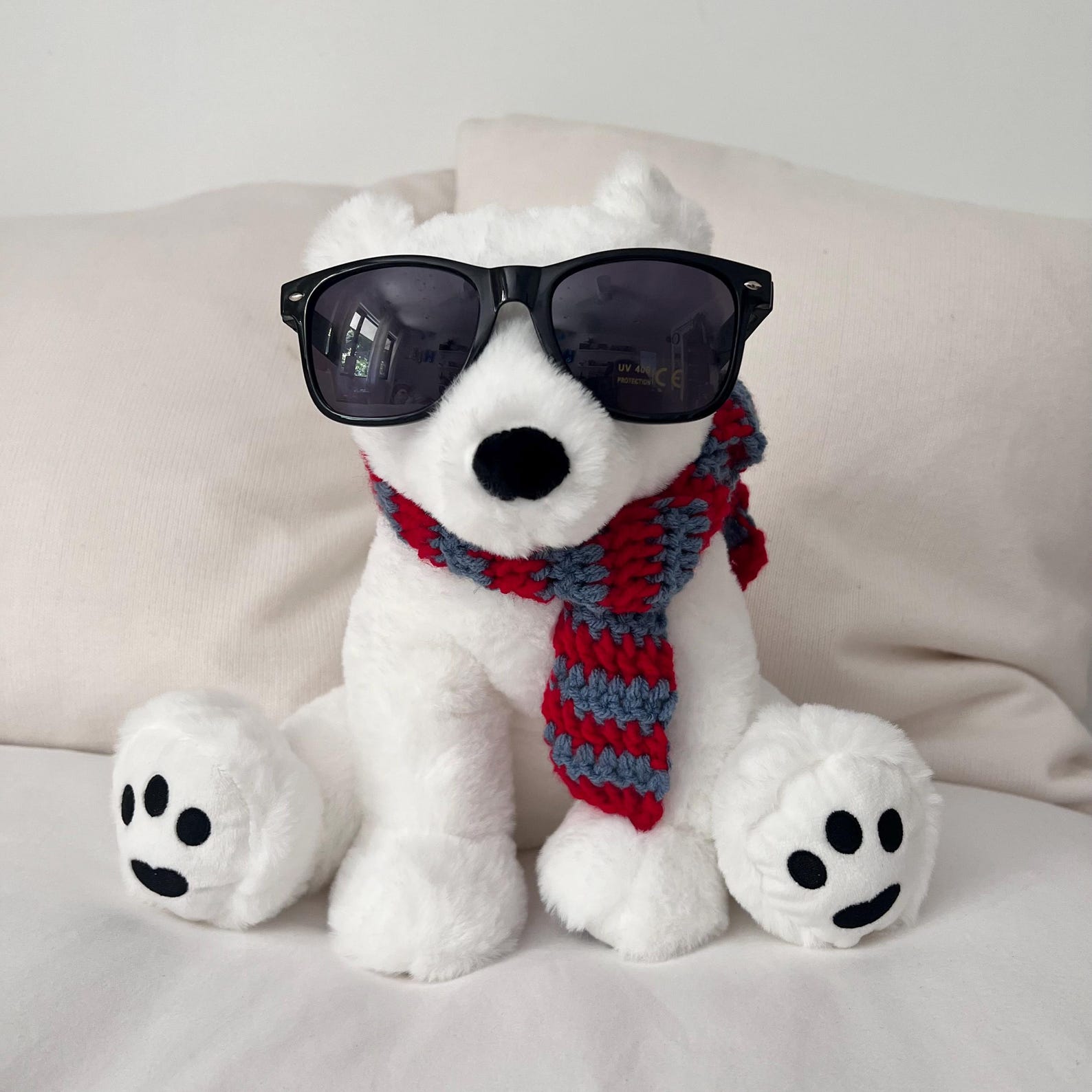 A plush polar facing the camera wearing oversized black sunglasses and a red and grey striped crocheted scarf.