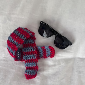 Peut inclure: Écharpe crochetée à rayures rouges et bleues, accompagnée de lunettes de soleil noires. L'écharpe est faite à la main avec un motif rayé. Les lunettes de soleil ont une monture rectangulaire classique.