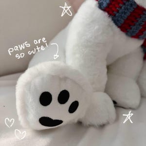 K&ouml;nnte beinhalten: Wei&szlig;er Pl&uuml;sch-Eisb&auml;r mit einem rot-blau gestreiften Schal. Der B&auml;r hat schwarze Pfoten und sitzt auf einer wei&szlig;en Oberfl&auml;che.