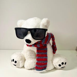 K&ouml;nnte beinhalten: Ein wei&szlig;er Pl&uuml;sch-Eisb&auml;r mit schwarzer Sonnenbrille und einem rot-blau gestreiften Schal.