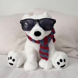 Könnte beinhalten: Ein weißes Eisbär-Stofftier mit schwarzer Nase und schwarzen Tatzen. Der Bär trägt eine schwarze Kunststoff-Sonnenbrille und einen grob gestrickten Schal mit abwechselnden horizontalen Streifen in den Farben Dunkelrot und Schieferblau.
