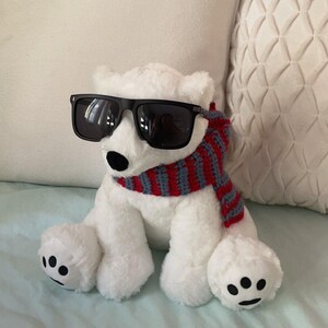 K&ouml;nnte beinhalten: Ein wei&szlig;er Pl&uuml;sch-Eisb&auml;r, der eine schwarze Sonnenbrille und einen rot-grauen gestreiften Schal tr&auml;gt. Der B&auml;r sitzt auf einer wei&szlig;en und blauen Oberfl&auml;che.