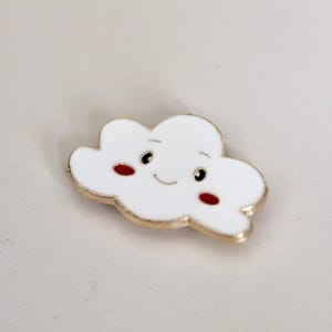 May include: A white enamel pin shaped like a cloud with a smiling face and red cheeks. The pin has a gold outline.