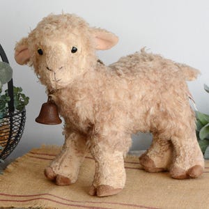 Puede incluir: Un cordero de peluche beige con una campana alrededor del cuello. El cordero tiene un pelaje suave y texturizado, orejas pequeñas y ojos oscuros. Se encuentra sobre una superficie beige con rayas rojas, y una cesta negra con vegetación es visible en el fondo.