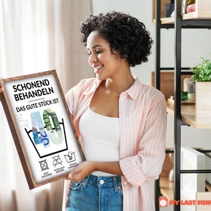 May include: A framed print with a white background and black text that reads "Schonend Behandeln Das Gute St&uuml;ck Ist 40". The number 40 is made of euro bills. There are four symbols below the number 40, representing laundry care instructions. The print is in a brown frame.