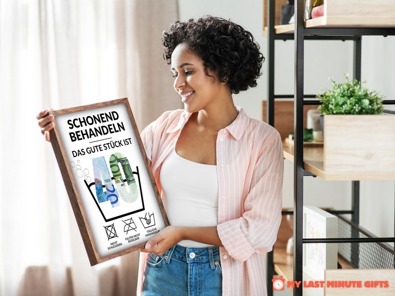 May include: A framed print with a white background and black text that reads "Schonend Behandeln Das Gute St&uuml;ck Ist 40". The number 40 is made of euro bills. There are four symbols below the number 40, representing laundry care instructions. The print is in a brown frame.