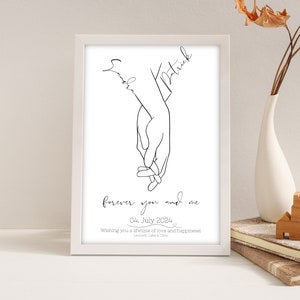 Könnte beinhalten: Ein weißer gerahmter Druck mit einer schwarzen Linienzeichnung von zwei Händen, die sich halten. Die Namen "Emily" und "Patrick" sind über den Händen geschrieben. Der Text "forever you and me" ist unter den Händen geschrieben, zusammen mit dem Datum "04. July 2024". Der Text "Wishing you a lifetime of love and happiness" ist unter dem Datum geschrieben.