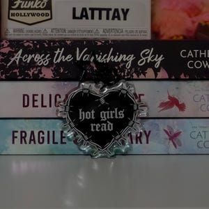 May include: A clear acrylic heart-shaped keychain featuring a black heart graphic wrapped in barbed wire. The text "hot girls read" is printed in a white gothic-style font across the center of the heart. A black star-shaped metal clasp is attached.