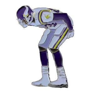 Op de afbeelding: Een zilveren metalen pin van een voetballer die een paars en wit uniform draagt met een gele streep. De speler buigt zich voorover met zijn hoofd naar beneden.