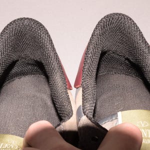 Puede incluir: Primer plano del interior de dos zapatillas deportivas. Los zapatos tienen un forro de malla gris oscuro y un acento rojo en el tal&oacute;n. El &aacute;rea de la leng&uuml;eta es visible, con una etiqueta que dice "Valentino".
