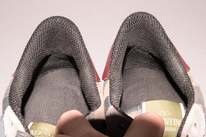 Puede incluir: Primer plano del interior de dos zapatillas deportivas. Los zapatos tienen un forro de malla gris oscuro y un acento rojo en el tal&oacute;n. El &aacute;rea de la leng&uuml;eta es visible, con una etiqueta que dice "Valentino".