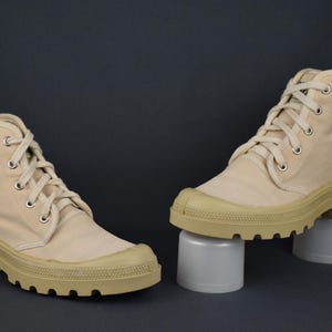 Puede incluir: Un par de botas altas de lona beige con suelas gruesas blancas. Las botas tienen cordones y est&aacute;n sobre una superficie blanca.