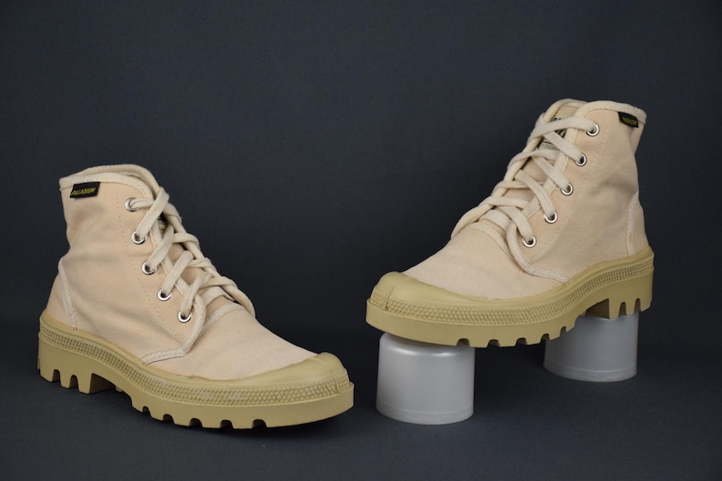 Puede incluir: Un par de botas altas de lona beige con suelas gruesas blancas. Las botas tienen cordones y est&aacute;n sobre una superficie blanca.