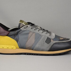 Puede incluir: Una zapatilla gris, negra y amarilla con un estampado de camuflaje. El zapato presenta una puntera negra, detalles de ante gris y un tal&oacute;n amarillo con detalles tachonados. La suela es de color beige y los cordones son grises.