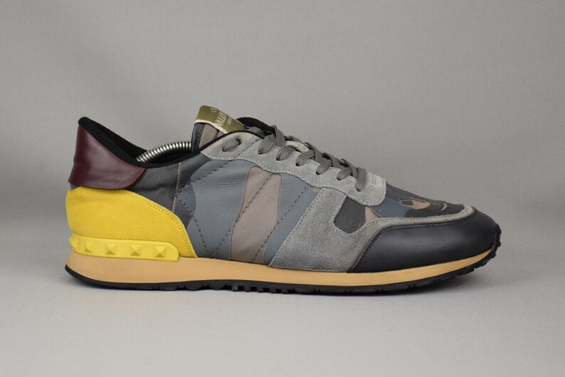 Puede incluir: Una zapatilla gris, negra y amarilla con un estampado de camuflaje. El zapato presenta una puntera negra, detalles de ante gris y un tal&oacute;n amarillo con detalles tachonados. La suela es de color beige y los cordones son grises.