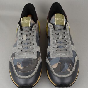 Puede incluir: Par de zapatillas deportivas Valentino Garavani. Los zapatos presentan un estampado de camuflaje en tonos grises, azules y marrones. Tienen detalles de ante gris, ribetes de cuero negro y una suela amarilla. El logotipo de Valentino Garavani aparece en la leng&uuml;eta.