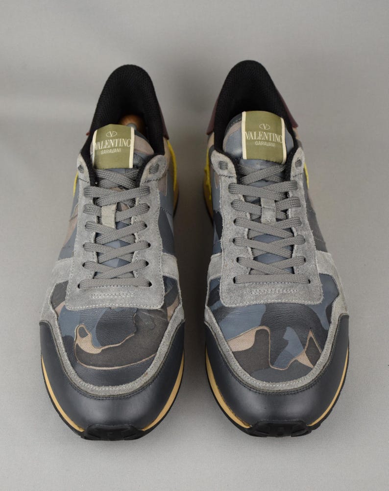 Puede incluir: Par de zapatillas deportivas Valentino Garavani. Los zapatos presentan un estampado de camuflaje en tonos grises, azules y marrones. Tienen detalles de ante gris, ribetes de cuero negro y una suela amarilla. El logotipo de Valentino Garavani aparece en la leng&uuml;eta.