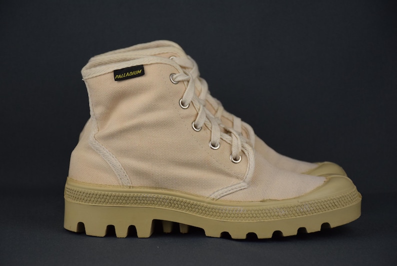 Puede incluir: Un par de botas altas de lona beige con una suela gruesa y texturizada. Las botas tienen cierre de cordones y una etiqueta amarilla "PALLADIUM" en el lateral.