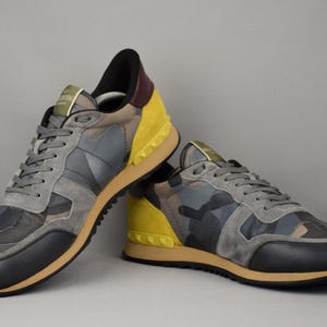 Puede incluir: Un par de zapatillas deportivas con un estampado de camuflaje en tonos grises, negros y marrones. Los zapatos tienen detalles amarillos en los laterales y un tal&oacute;n negro. Las suelas son color canela y los cordones grises.