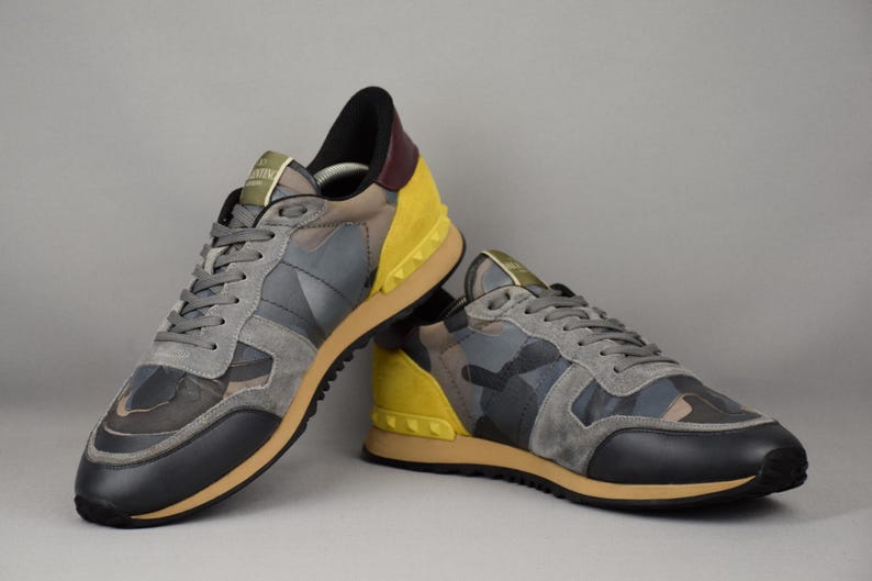 Puede incluir: Un par de zapatillas deportivas con un estampado de camuflaje en tonos grises, negros y marrones. Los zapatos tienen detalles amarillos en los laterales y un tal&oacute;n negro. Las suelas son color canela y los cordones grises.