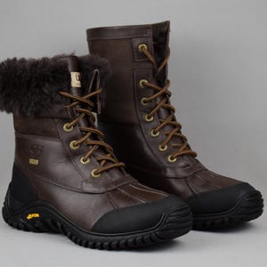 May include: A pair of brown leather winter boots with dark brown fur trim at the top. The boots have brown laces, a black rubber sole, and the UGG logo. The boots are designed for cold weather and outdoor activities.