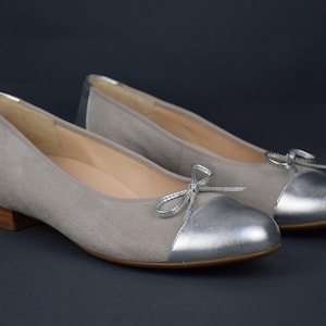 以下が含まれることがあります： シルバーのつま先とリボンが付いたグレーのスエードのバレエシューズのペア。フラットはローヒールです。