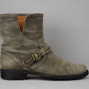 May include: A gray suede ankle boot with a low heel. The boot features a buckle strap with gold-colored hardware and decorative studs. The top of the boot has a V-shaped cutout. The sole is black.