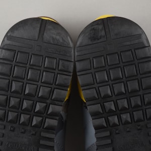 Puede incluir: Primer plano de las suelas de dos zapatillas deportivas. Las suelas son negras con un patr&oacute;n texturizado en forma de rejilla y peque&ntilde;os tachuelas en forma de pir&aacute;mide. Los talones tienen un acento amarillo. La marca "Valentino" es visible.