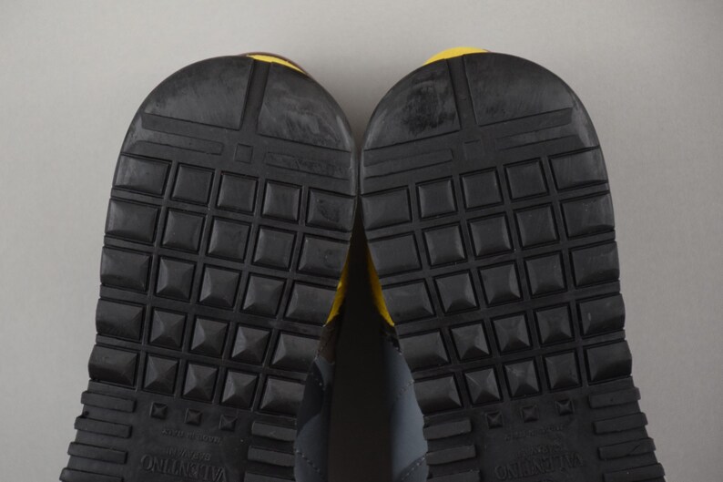 Puede incluir: Primer plano de las suelas de dos zapatillas deportivas. Las suelas son negras con un patr&oacute;n texturizado en forma de rejilla y peque&ntilde;os tachuelas en forma de pir&aacute;mide. Los talones tienen un acento amarillo. La marca "Valentino" es visible.