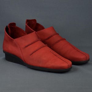 May include: A pair of red slip-on shoes featuring a matte finish, a low wedge heel, and a black sole. The upper design includes two horizontal stitched pleats across the vamp and a vertical seam running down the center of the rounded toe box.