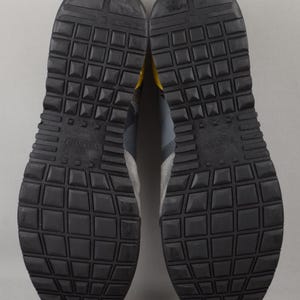 Puede incluir: Un par de zapatillas grises y negras con suela de goma texturizada. Las suelas son negras con un patr&oacute;n de rejilla. Las zapatillas tienen paneles de ante y cuero grises con detalles en amarillo.