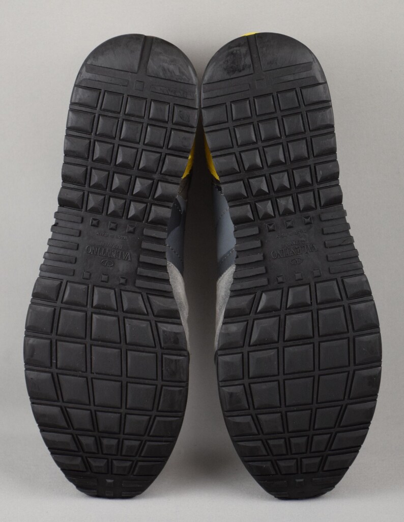 Puede incluir: Un par de zapatillas grises y negras con suela de goma texturizada. Las suelas son negras con un patr&oacute;n de rejilla. Las zapatillas tienen paneles de ante y cuero grises con detalles en amarillo.