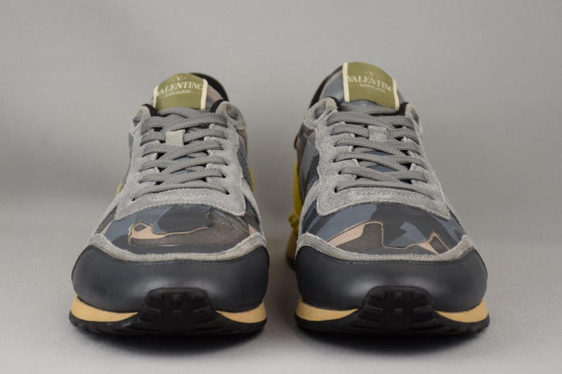 Puede incluir: Par de zapatillas grises y camuflaje con suelas negras y detalles beige. Las zapatillas tienen cordones grises y el nombre de la marca es visible en la leng&uuml;eta. Los zapatos est&aacute;n dise&ntilde;ados para uso casual.