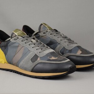Puede incluir: Un par de zapatillas deportivas con un estampado de camuflaje en tonos grises, azules y marrones. Los zapatos tienen detalles en negro y amarillo, cordones grises y una suela color canela. Las zapatillas se muestran sobre un fondo gris.