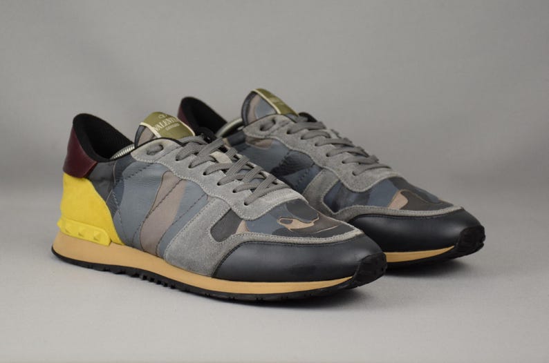 Puede incluir: Un par de zapatillas deportivas con un estampado de camuflaje en tonos grises, azules y marrones. Los zapatos tienen detalles en negro y amarillo, cordones grises y una suela color canela. Las zapatillas se muestran sobre un fondo gris.