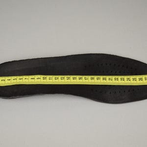 Puede incluir: Una plantilla de zapato negra con una cinta m&eacute;trica amarilla colocada encima. La plantilla mide aproximadamente 30 cm de largo. La plantilla tiene peque&ntilde;os orificios de ventilaci&oacute;n.