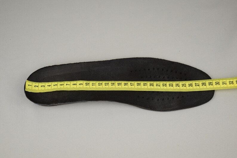 Puede incluir: Una plantilla de zapato negra con una cinta m&eacute;trica amarilla colocada encima. La plantilla mide aproximadamente 30 cm de largo. La plantilla tiene peque&ntilde;os orificios de ventilaci&oacute;n.
