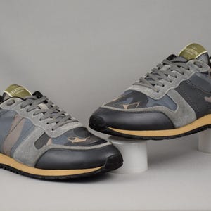 Puede incluir: Par de zapatillas grises, negras y de camuflaje con detalles en amarillo y burdeos. Las zapatillas tienen cordones grises y suela color canela. Los zapatos se exhiben sobre pedestales blancos sobre un fondo gris.