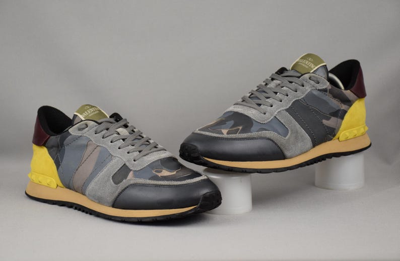 Puede incluir: Par de zapatillas grises, negras y de camuflaje con detalles en amarillo y burdeos. Las zapatillas tienen cordones grises y suela color canela. Los zapatos se exhiben sobre pedestales blancos sobre un fondo gris.