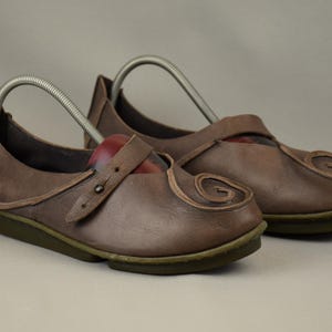May include: A pair of brown leather Mary Jane style shoes. The shoes have a strap with a button closure and a decorative swirl design on the toe. The soles are green.