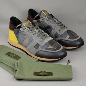 Puede incluir: Un par de zapatillas de camuflaje con detalles de ante gris y un panel de tal&oacute;n amarillo. Las zapatillas van acompa&ntilde;adas de bolsas de tela verdes para zapatos. Los zapatos tienen una puntera negra y una suela beis.