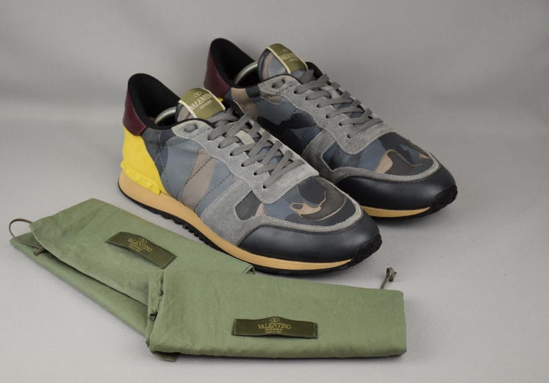 Puede incluir: Un par de zapatillas de camuflaje con detalles de ante gris y un panel de tal&oacute;n amarillo. Las zapatillas van acompa&ntilde;adas de bolsas de tela verdes para zapatos. Los zapatos tienen una puntera negra y una suela beis.