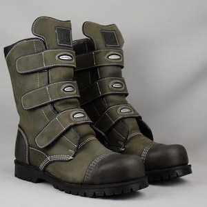 Könnte beinhalten: Ein Paar olivgrüne Lederstiefel mit mehreren verstellbaren Riemen und weißen Nähten. Die Stiefel haben ein robustes Design mit verstärkter Spitze und einer dicken, schwarzen Sohle. Die Stiefel haben oben die Buchstaben "UC". Höhe ca. 30 cm.