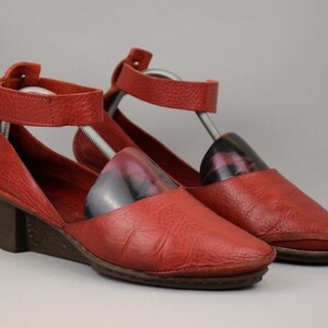 May include: A pair of red leather wedge sandals with ankle straps. The shoes have a textured finish and a dark brown wedge heel. The sandals are displayed with shoe trees inside.