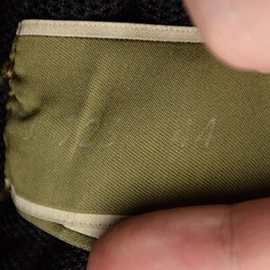 Puede incluir: Primer plano de una etiqueta de tela verde con un borde beige. La etiqueta est&aacute; parcialmente oscurecida por una mano. La tela tiene una apariencia texturizada. Texto blanco tenue visible en la etiqueta.